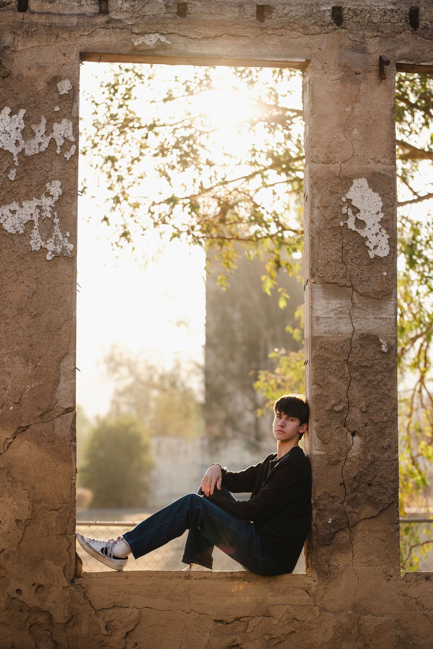 Young man in black outfit sitting in rustic stone window frame with sunlit trees behind him.