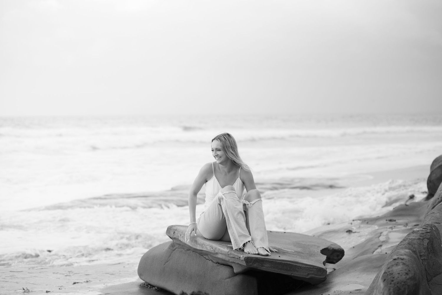 Black and white photo of a smiling woman sitting on rocks at a beach with ocean waves in the background.