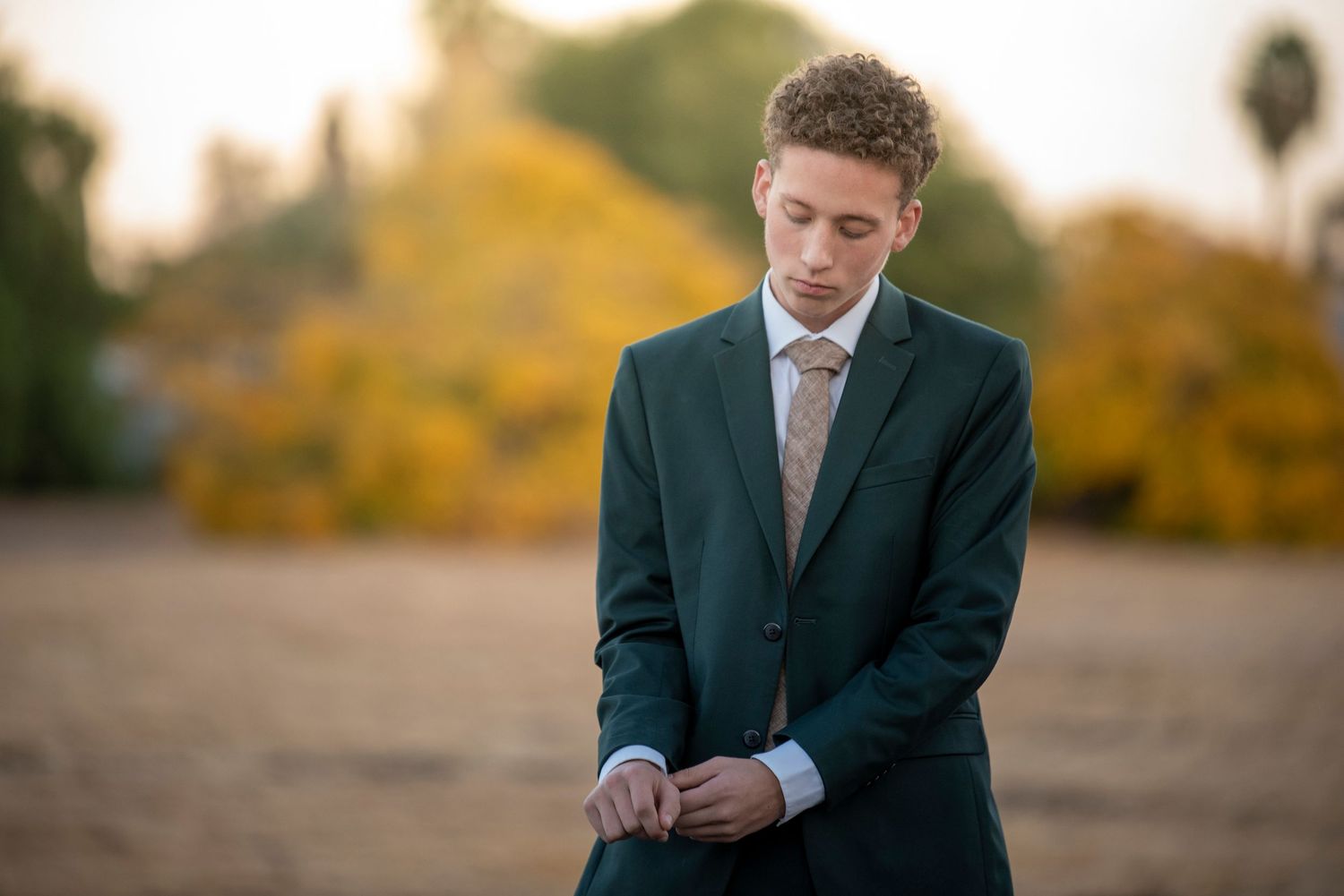 Young man in dark green suit adjusting jacket buttons while looking down in outdoor autumn setting.