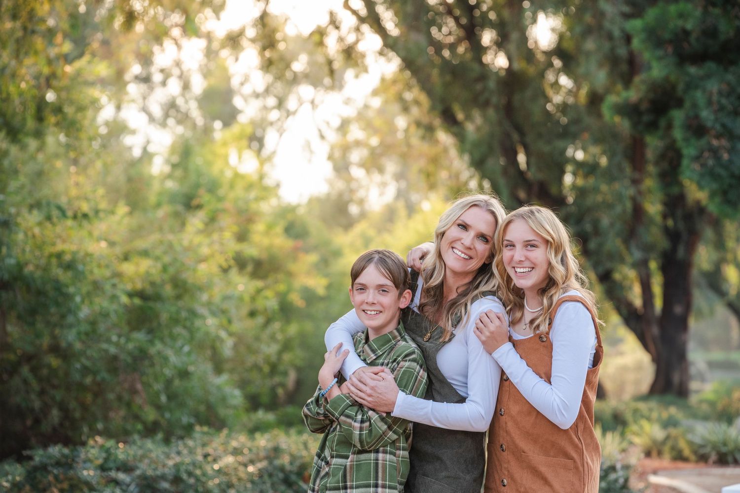 A smiling woman hugs two children outdoors among green trees in warm natural light.