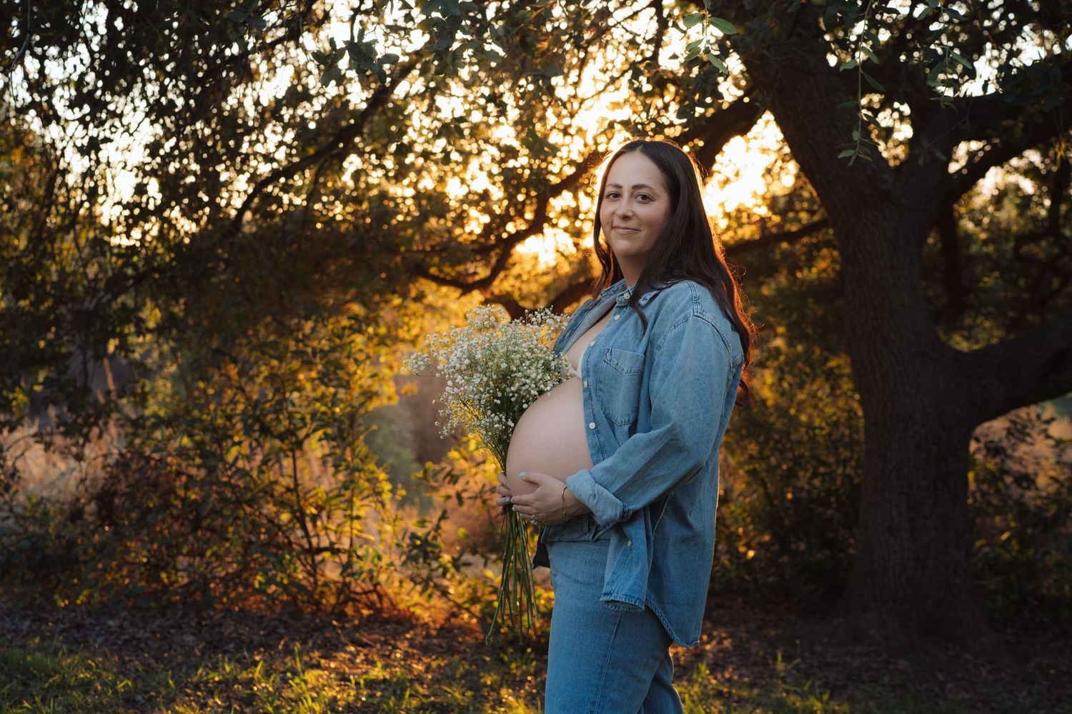 Smiling pregnant woman holding white wildflowers under a tree at golden sunset.