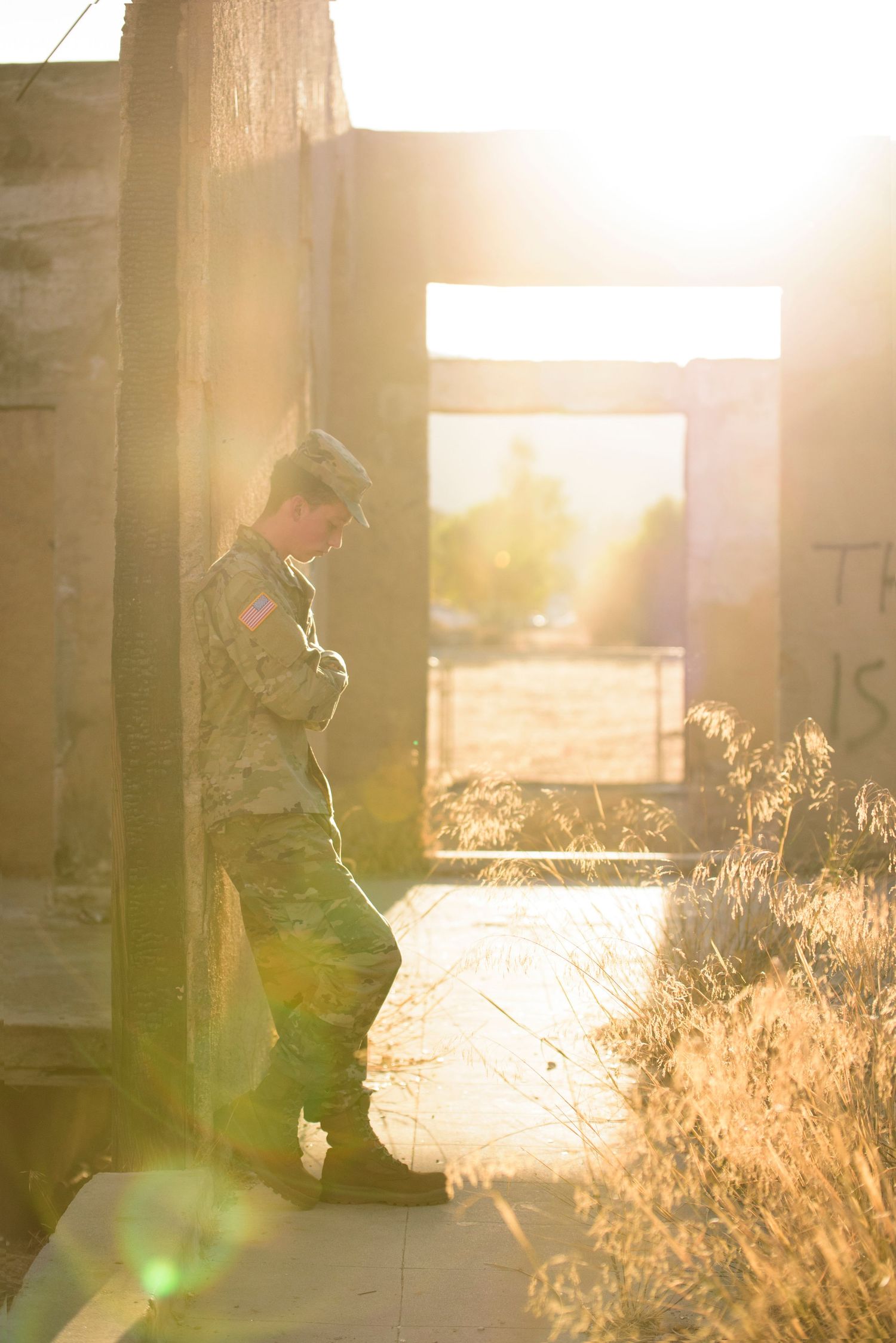 Female soldier in camouflage uniform leaning against concrete wall in golden sunlight at abandoned building.