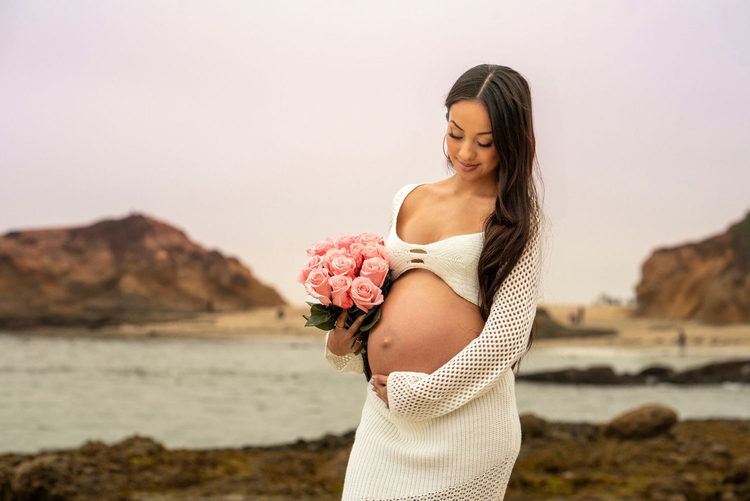 Pregnant woman in white dress holding pink roses bouquet at rocky beach, gazing down at her bare baby bump.