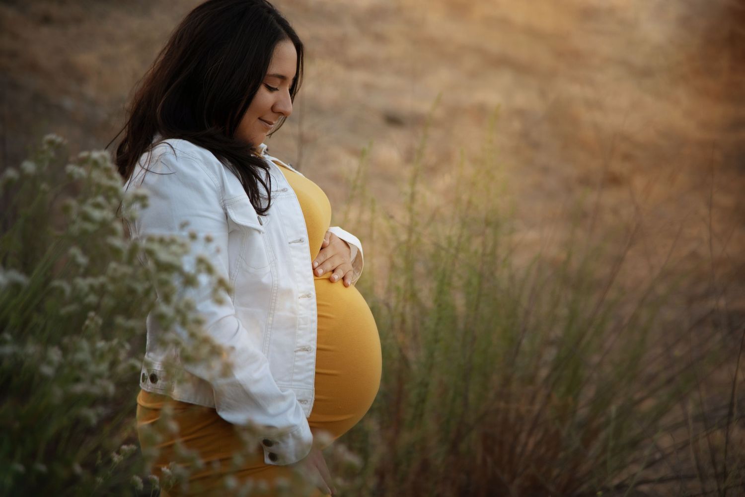 Smiling pregnant woman in yellow dress and white jacket cradling baby bump in sunlit field.