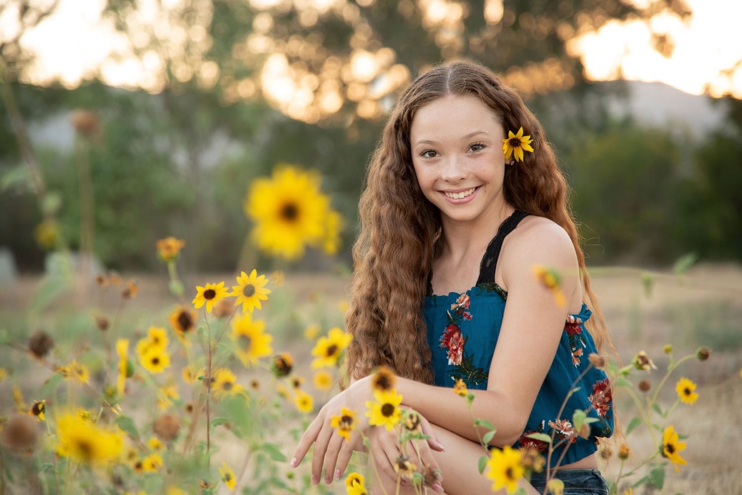 Smiling teenage girl with curly hair and yellow flowers posing in a sunflower field at golden hour.