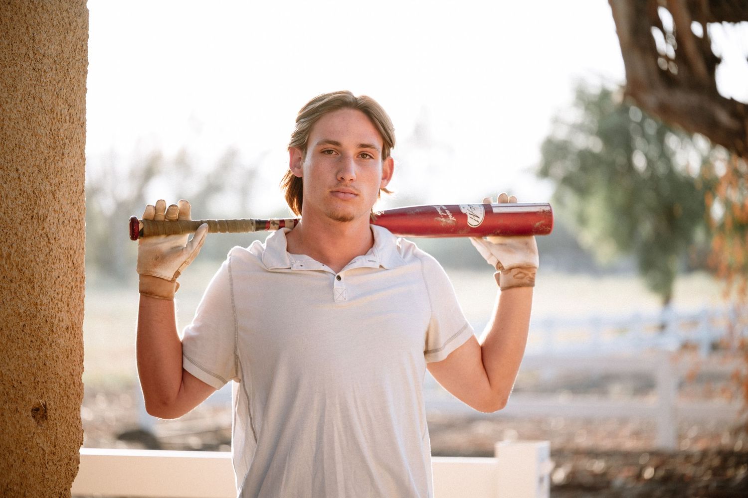 Young man in white polo shirt holds baseball bat over shoulder outdoors in warm sunlight.