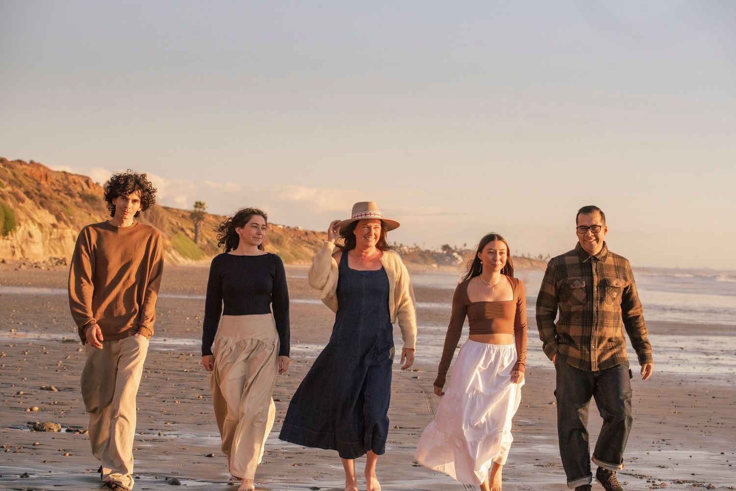 A family of five walks along a sunny beach with coastal cliffs in the background.