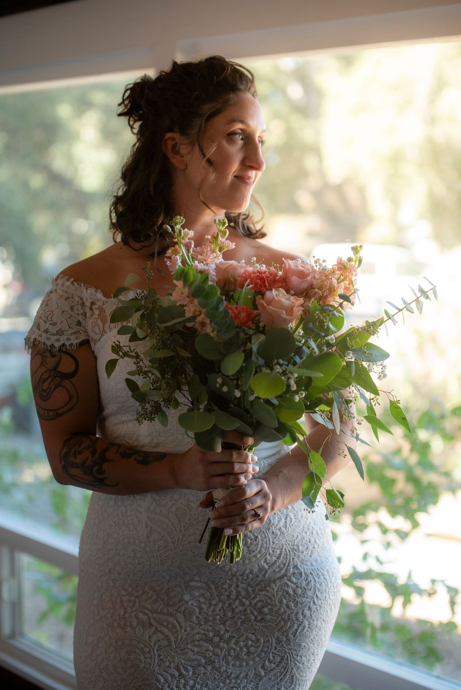 Tattooed bride in lace dress holding a pink and peach bouquet while gazing out a sunlit window.
