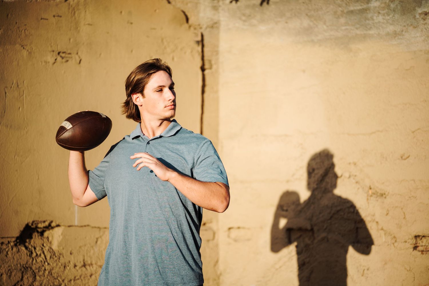 Young man in blue polo shirt throws football near golden wall with shadow visible behind him.