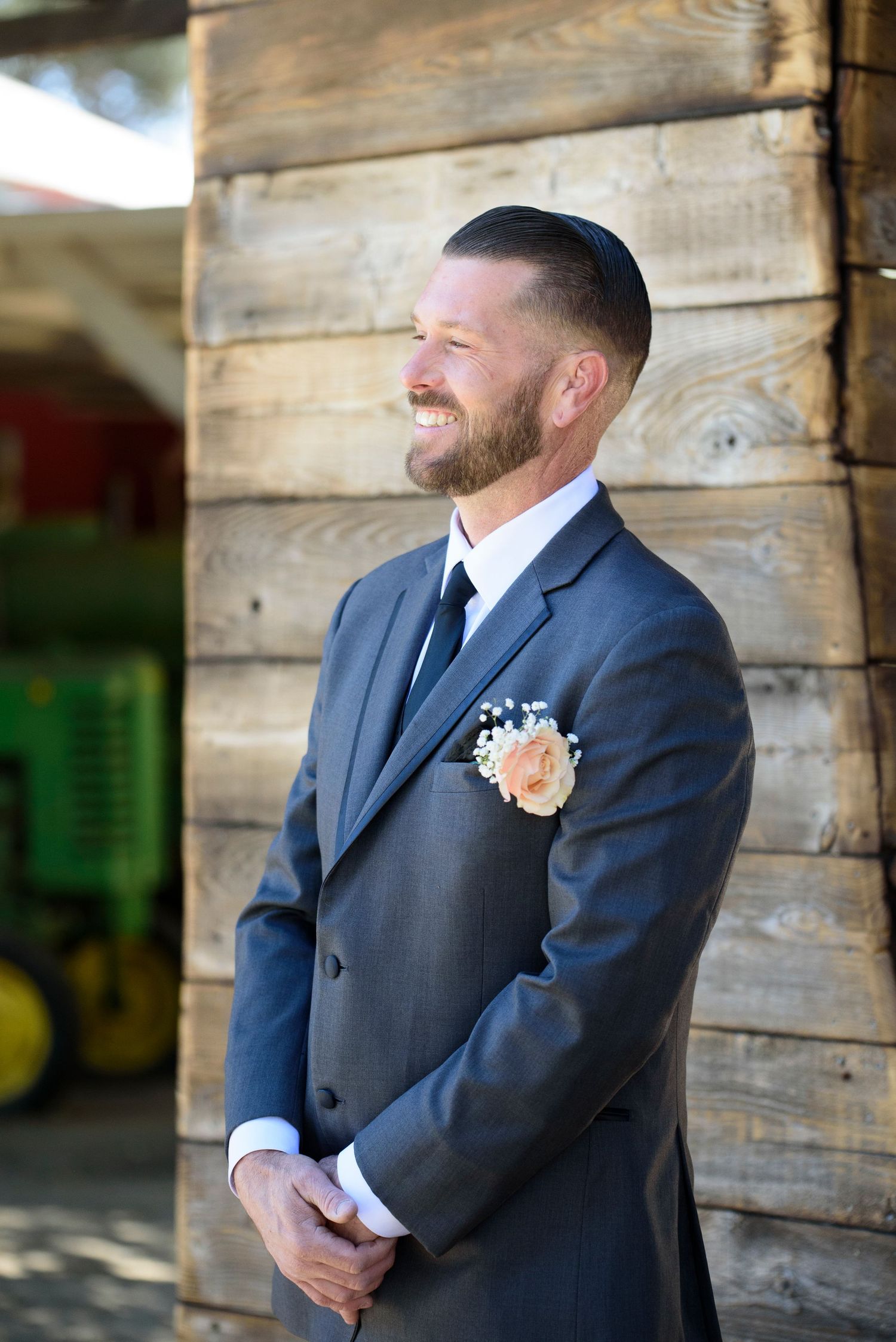Bearded groom in gray suit with boutonniere smiles while leaning against rustic wooden barn wall.