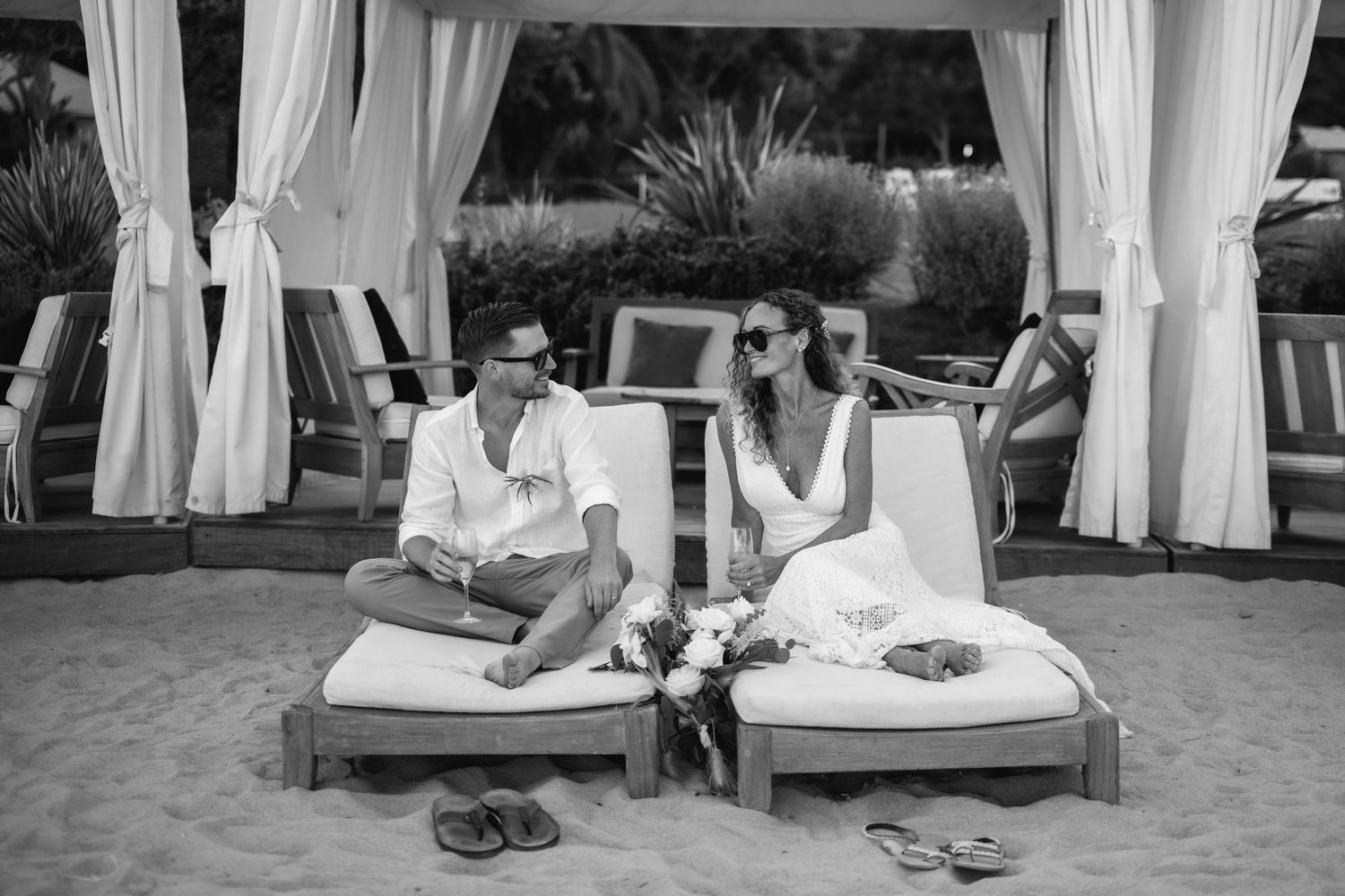 Black and white photo of a couple in wedding attire sitting on beach loungers, smiling at each other near cabanas.