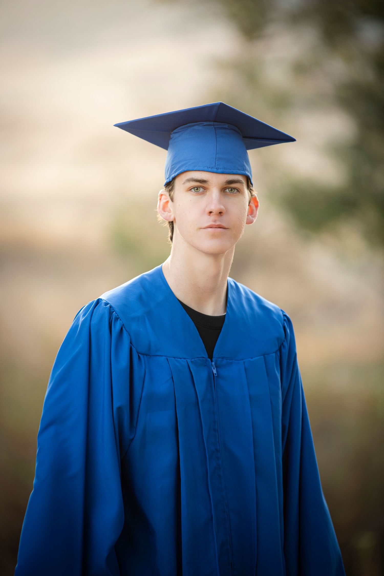 Young man wearing a blue graduation cap and gown posing outdoors with a soft blurred background.