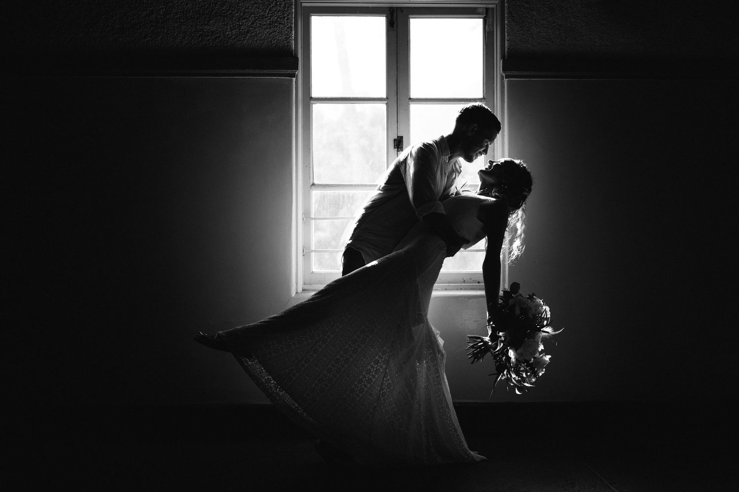 Black and white photo of a couple sharing a romantic dip kiss in front of a window on their wedding day.