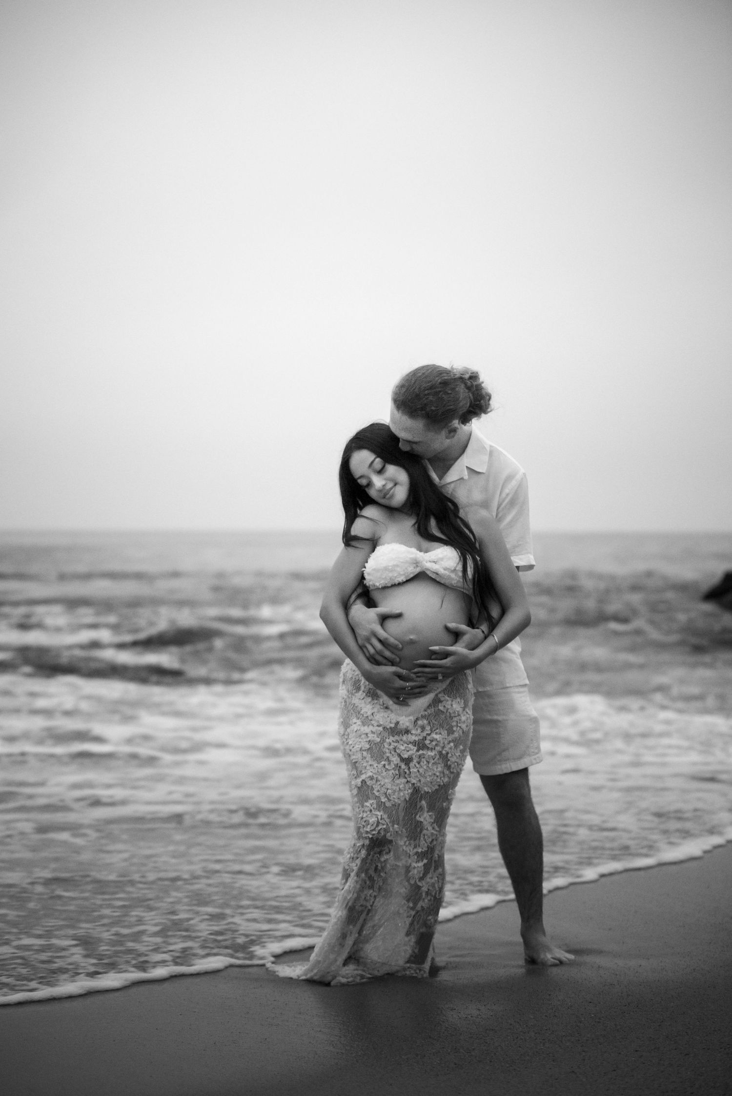 Couple sharing tender maternity moment on beach shoreline, man embracing pregnant woman in lace dress.