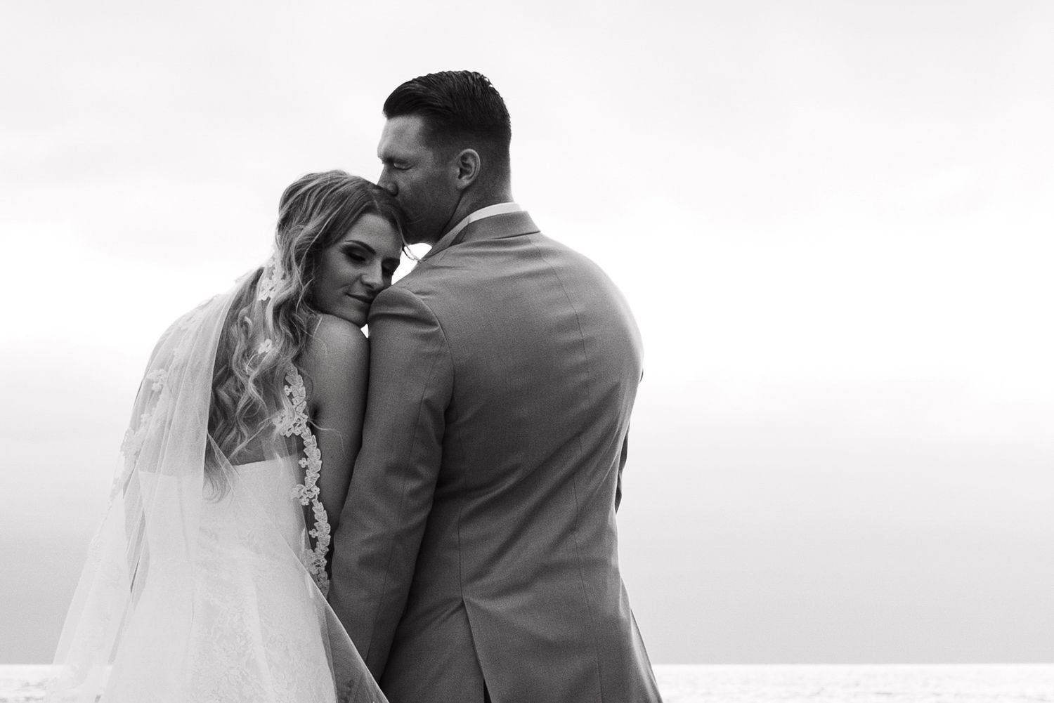 Black and white wedding photo of bride and groom embracing on beach, groom kissing bride's head.