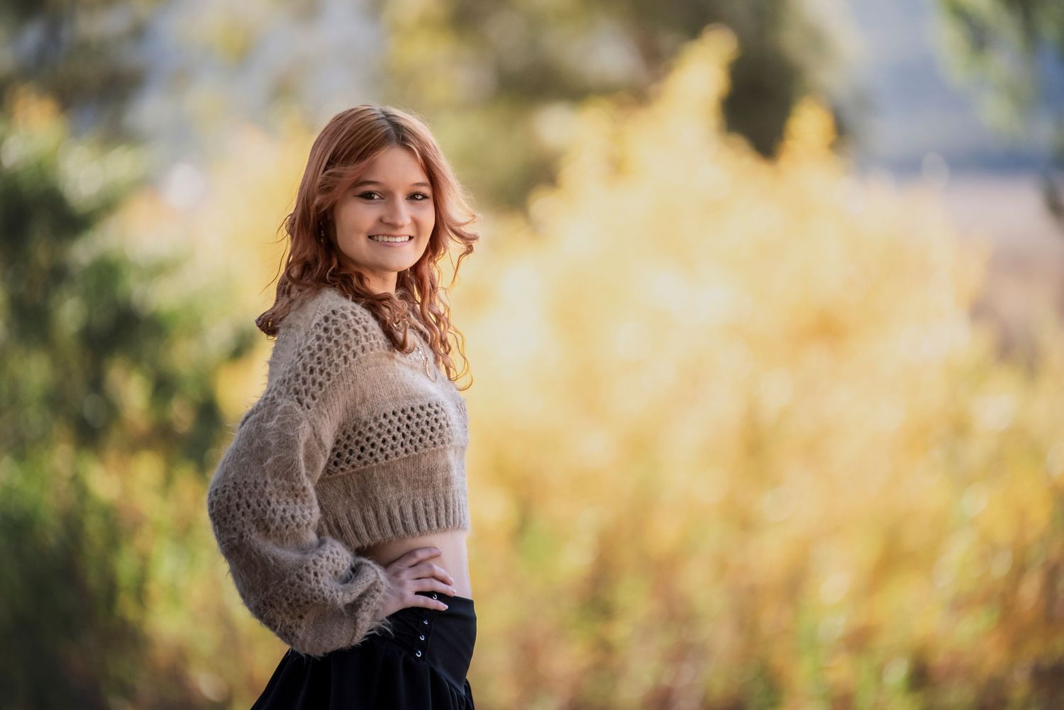 Smiling teenage girl in beige knit sweater posing outdoors with autumn foliage background.