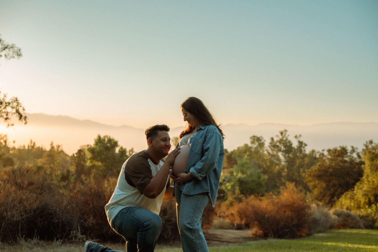 Man kneeling before pregnant partner at golden sunset in an outdoor field setting.