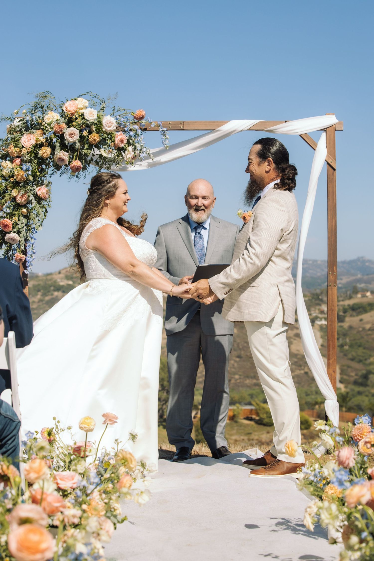 Bride and groom exchange vows under floral arch at outdoor hilltop wedding ceremony on sunny day.