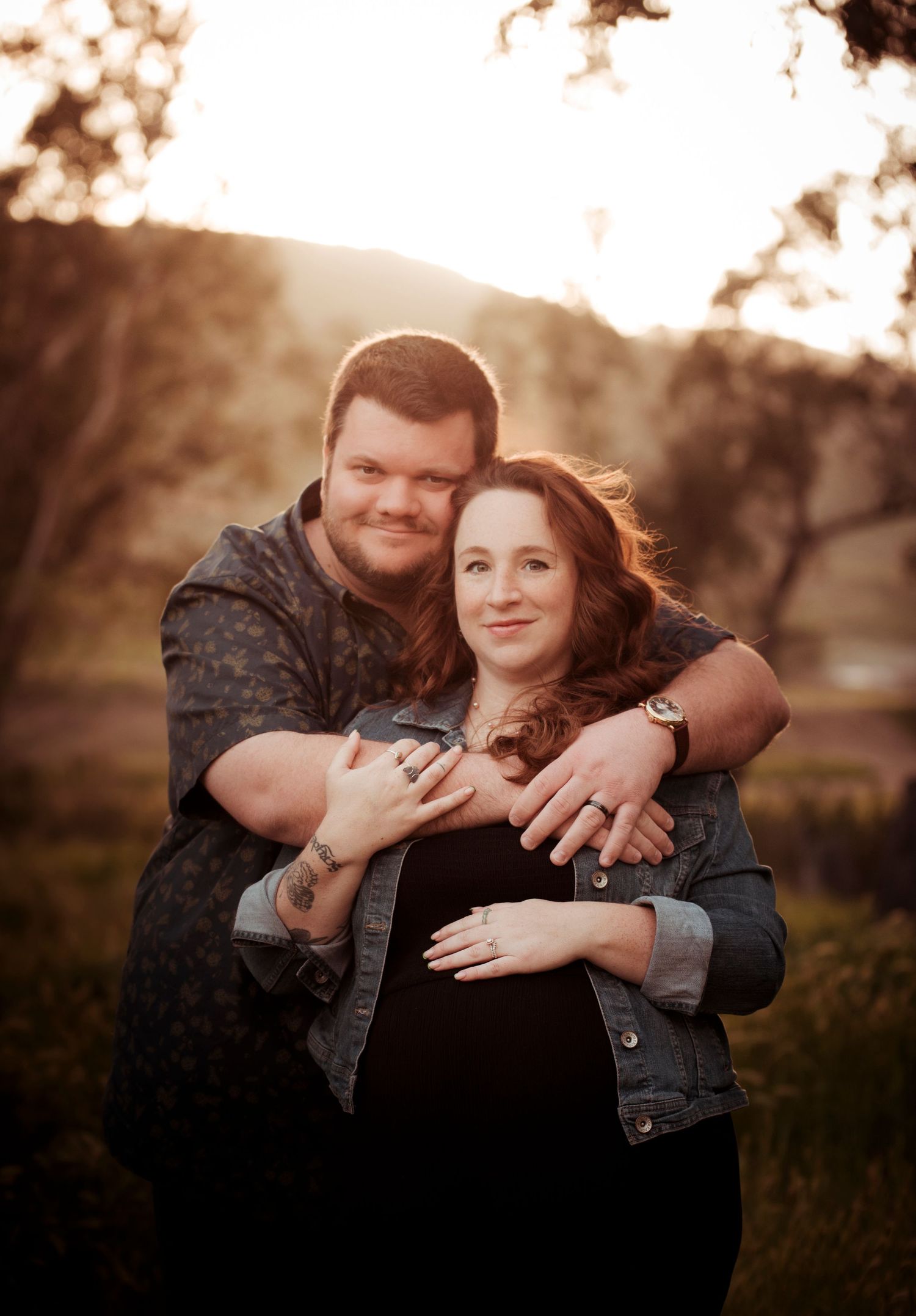Couple embracing warmly at sunset outdoors, woman's pregnant belly visible as partner wraps arms around her in a field.