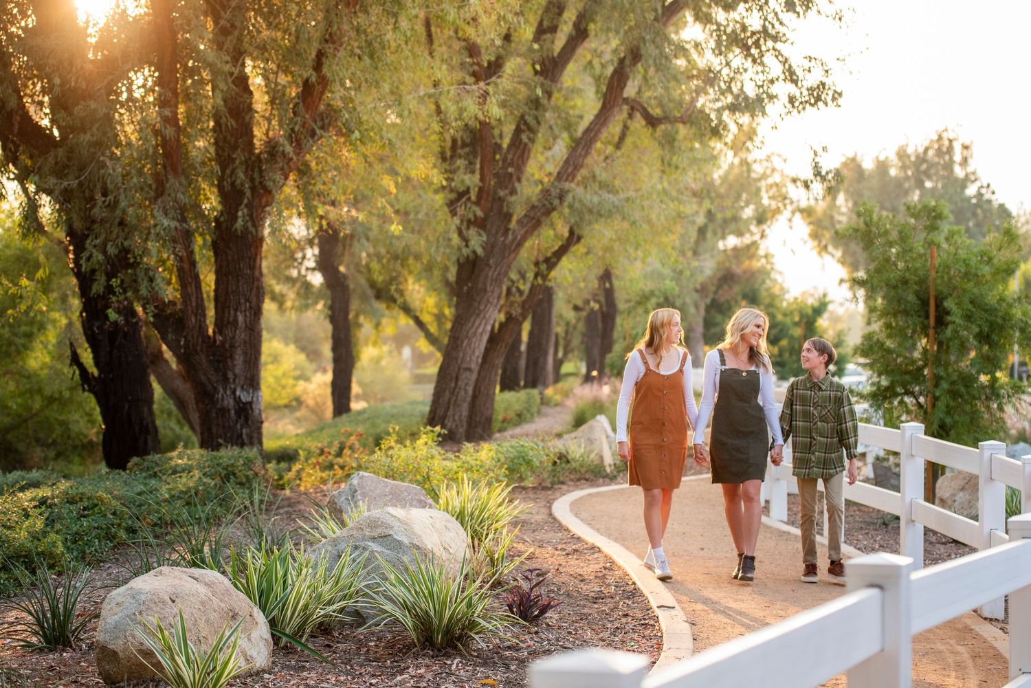 Three people walking along a sunlit garden path lined with trees and white fence.