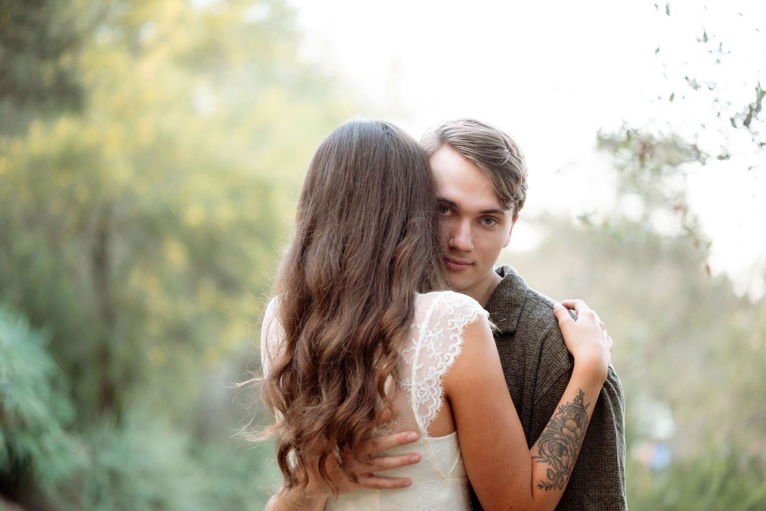 A couple embraces outdoors, the man with a tattoo gazing over the woman's shoulder amid soft green foliage.