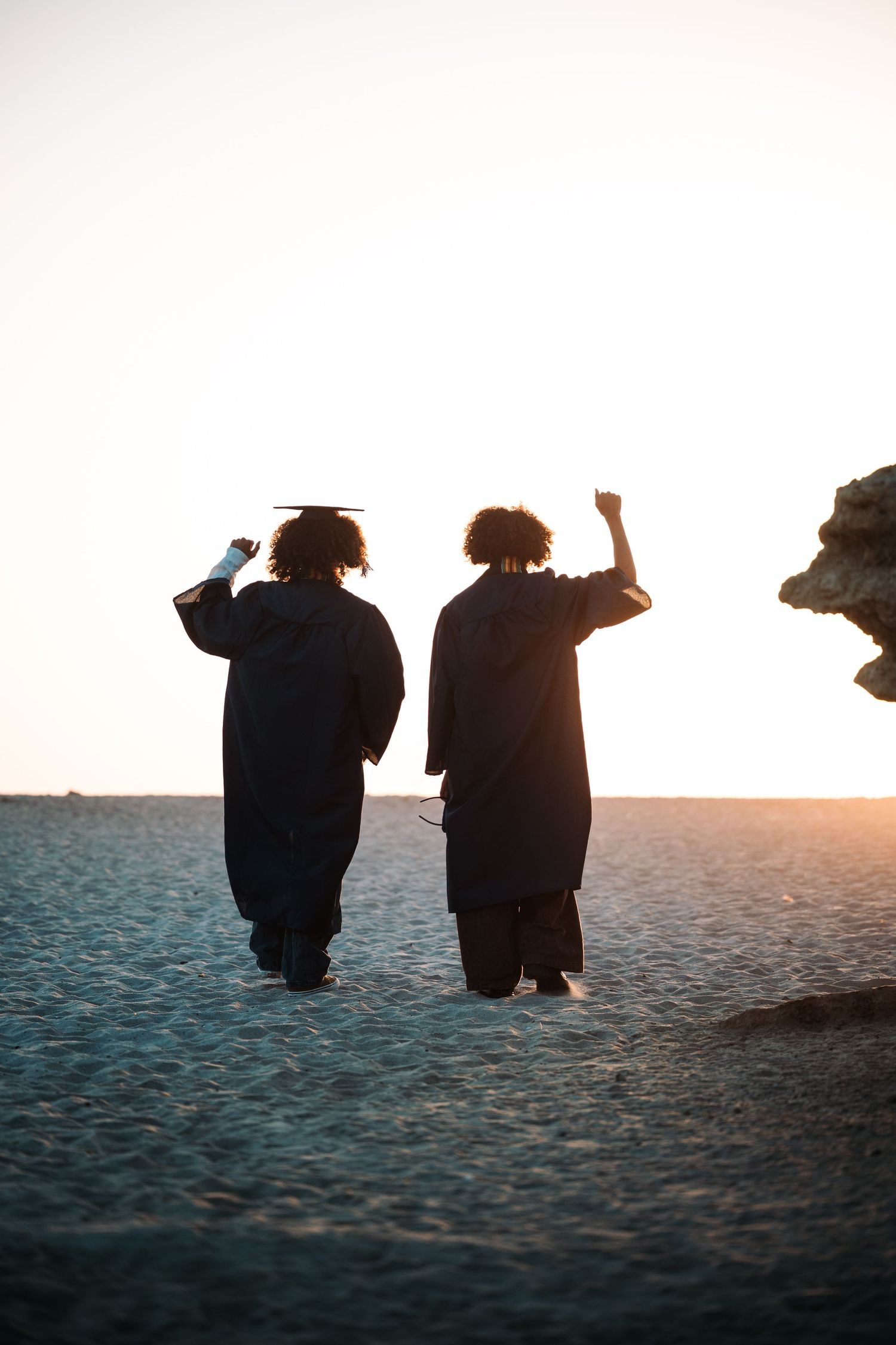 Two graduates in black robes celebrate at sunset on a beach, one raising fist triumphantly toward sky.