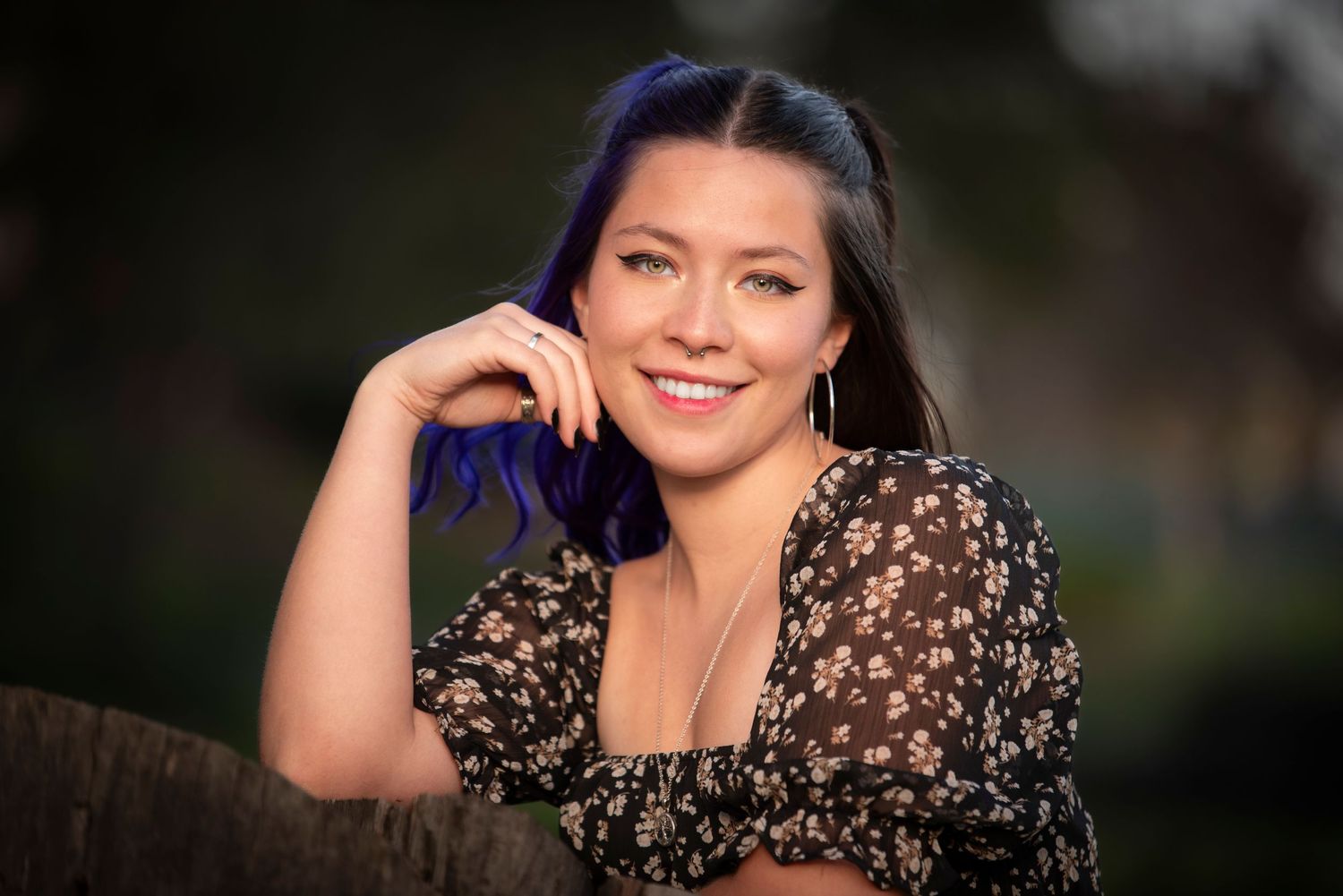 Smiling woman with blue hair wearing floral dress posing outdoors in soft light.