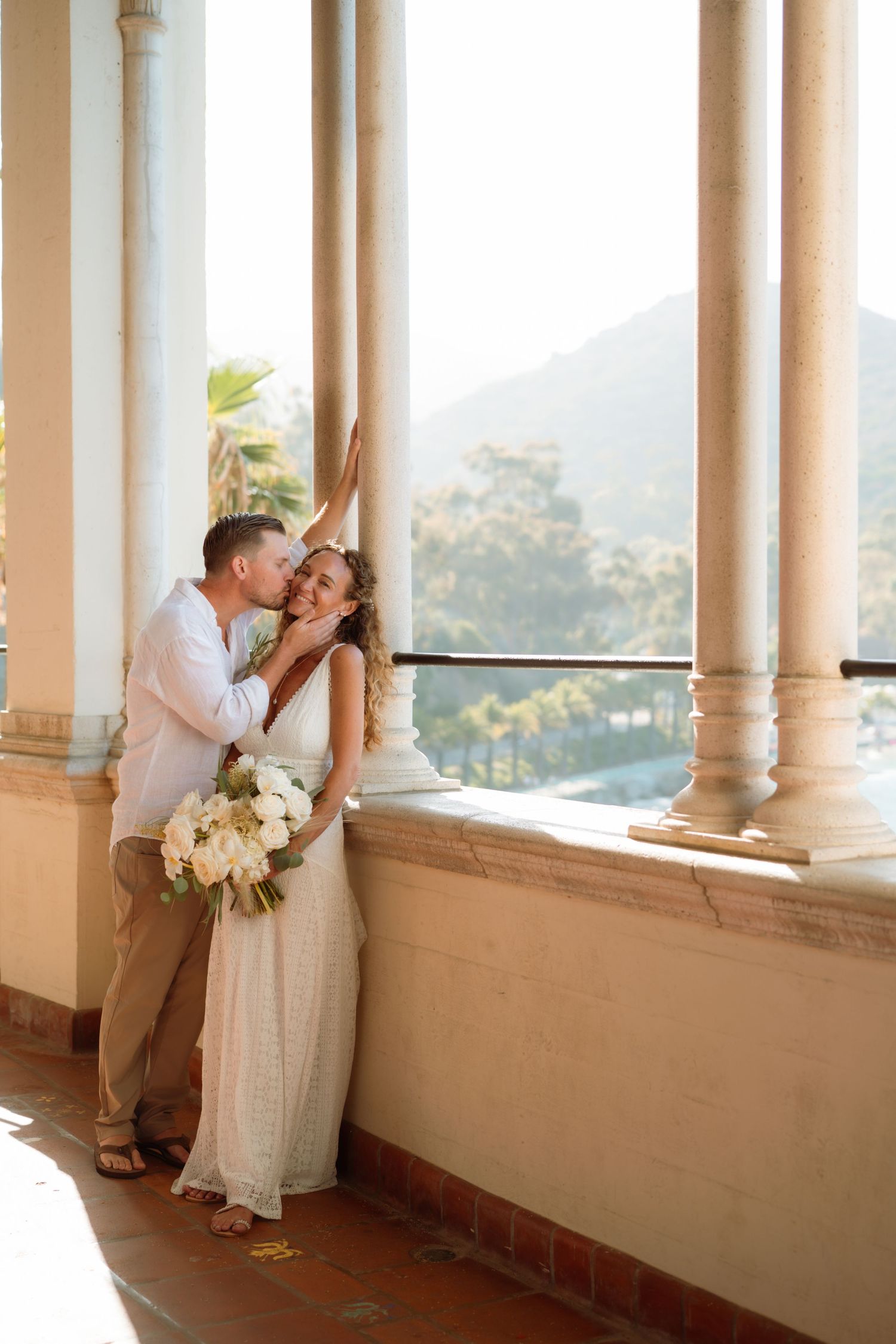 Bride and groom share a romantic kiss on a sunlit balcony with columns and mountain views.