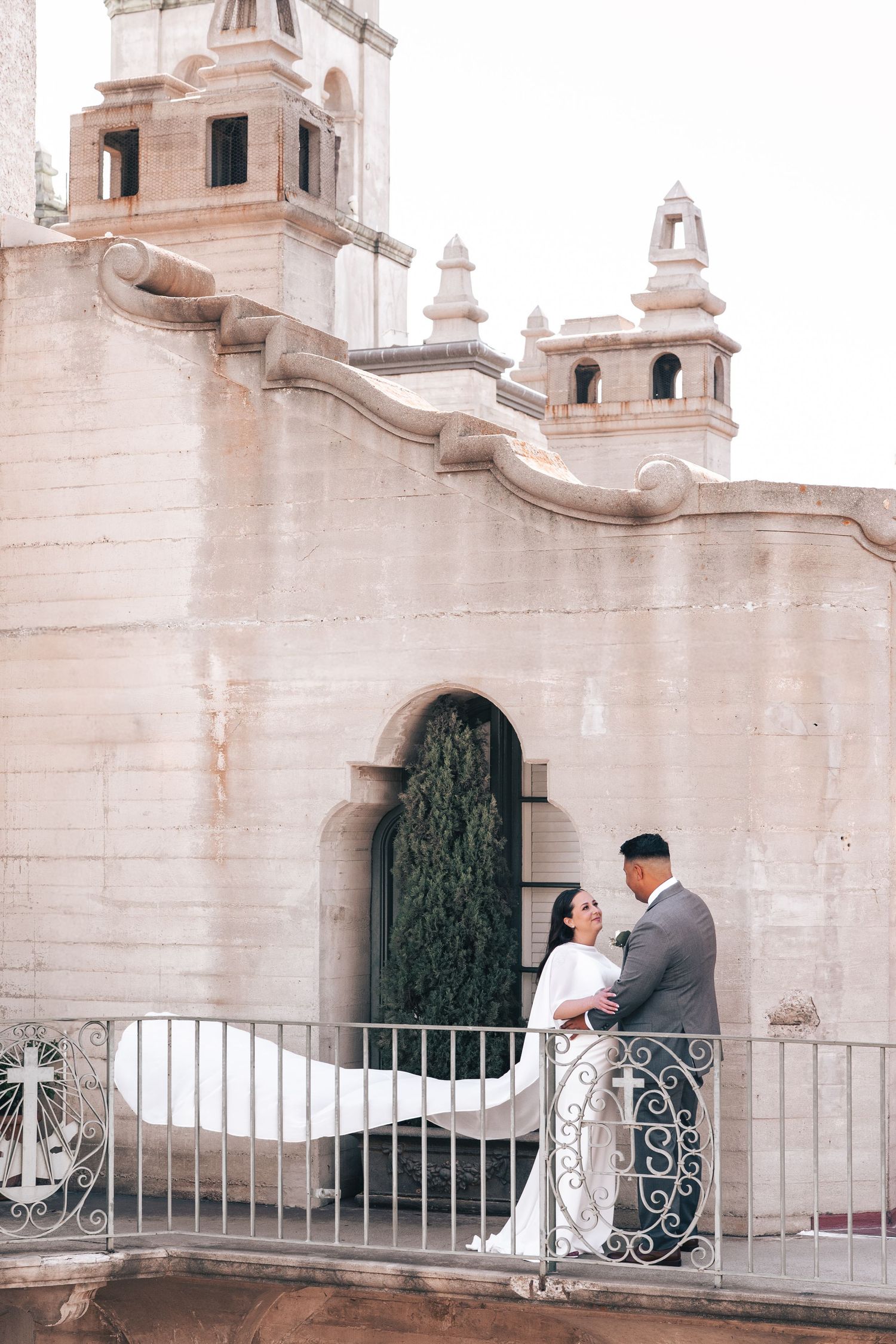Bride and groom embracing on a balcony beside a historic stone church exterior.