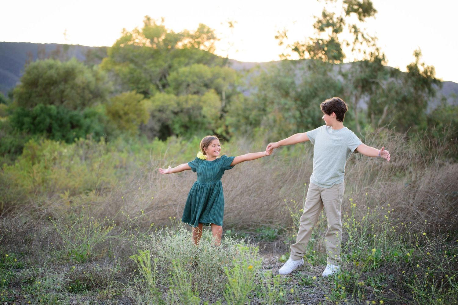 A young boy and girl hold hands with arms outstretched in a sunlit field of tall green grass.
