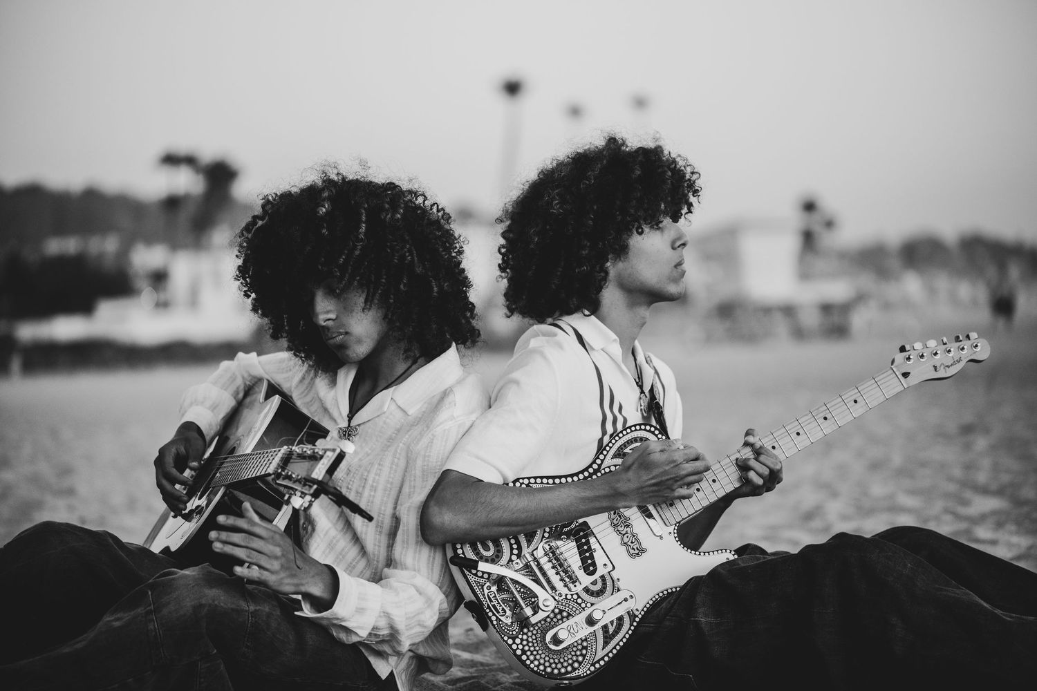 Two curly-haired musicians playing guitars back-to-back on a beach in black and white.