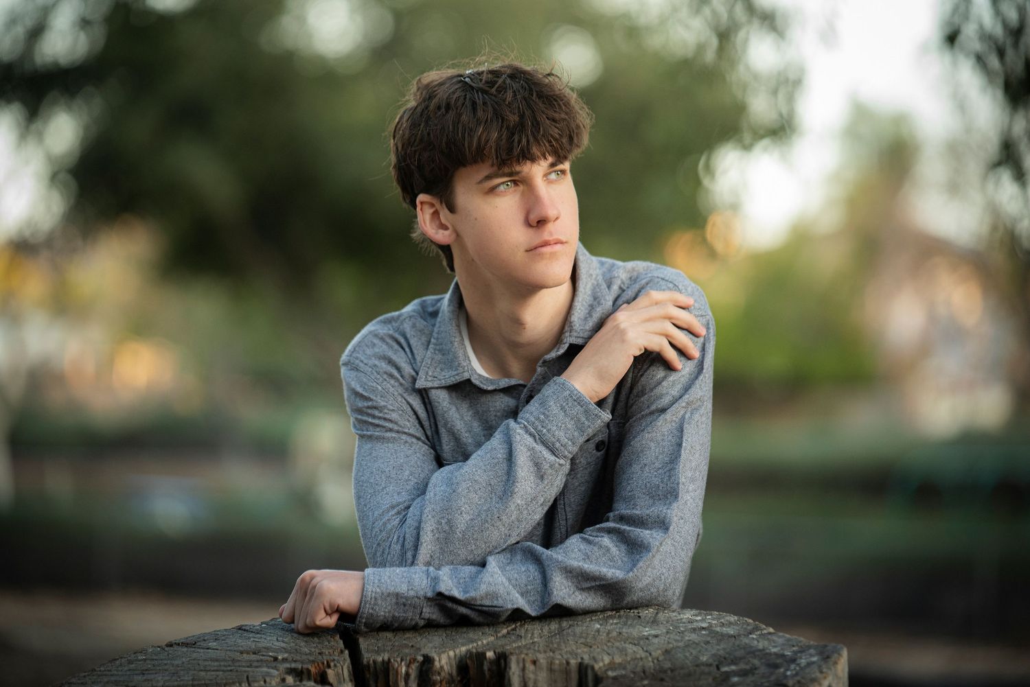 Young man in gray shirt resting crossed arms on a wooden post while gazing thoughtfully outdoors.