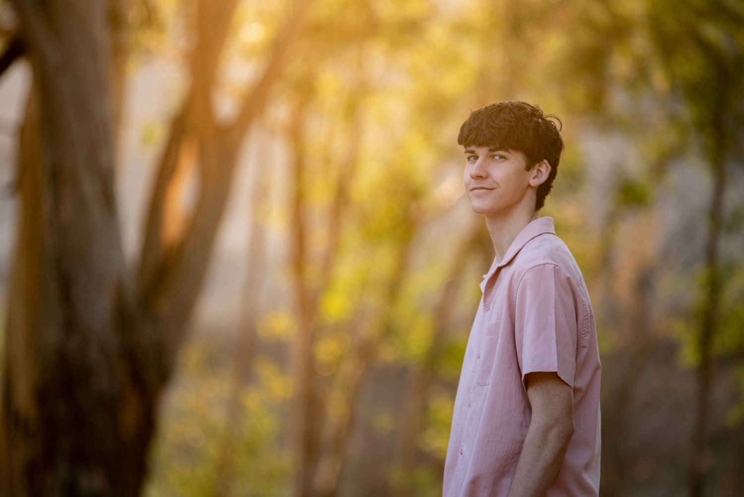 Young man in a pink polo shirt standing outdoors among trees in warm golden sunlight.