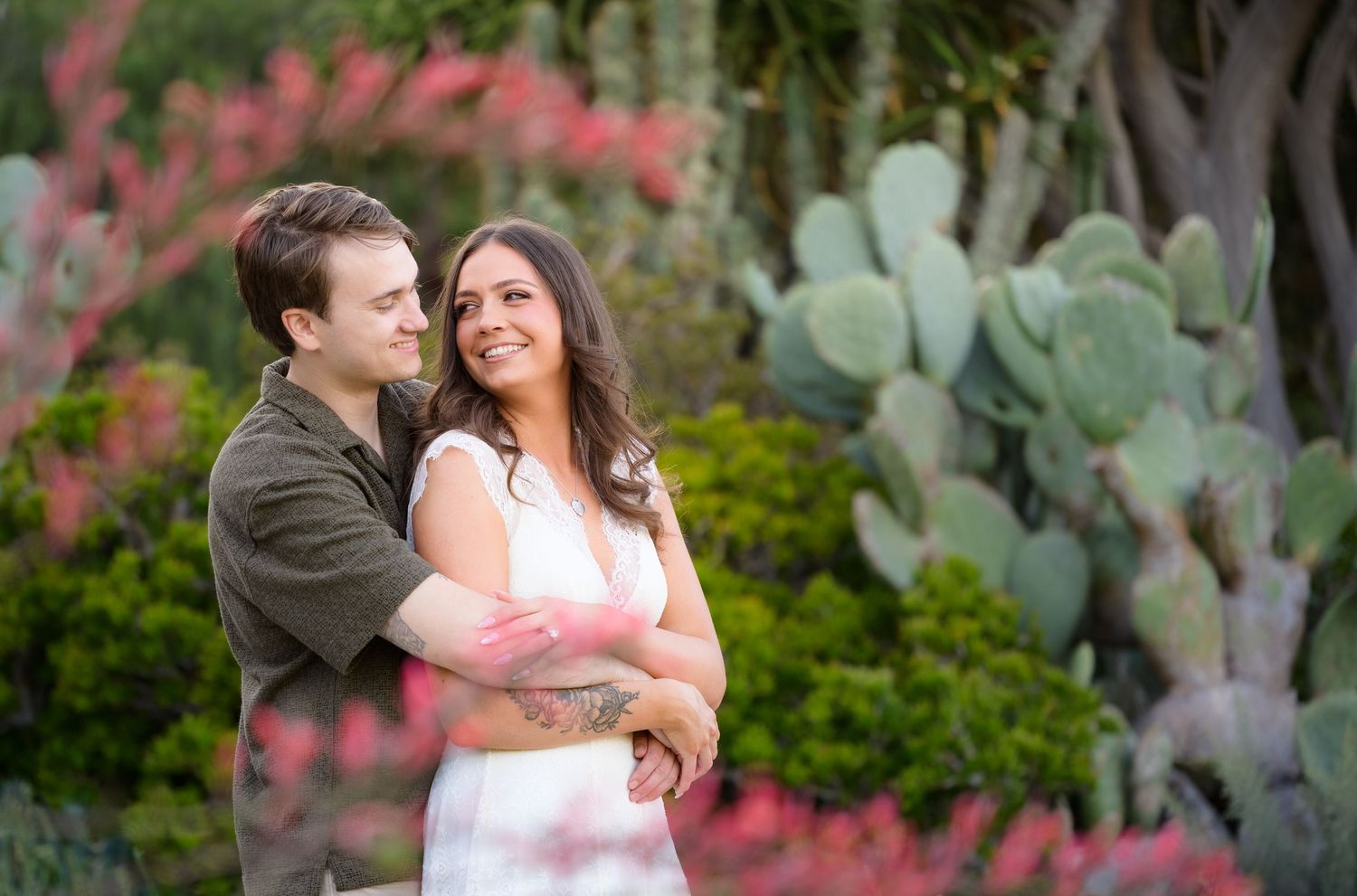 Couple embracing outdoors near cacti and pink flowers, smiling together in a romantic engagement photo session.