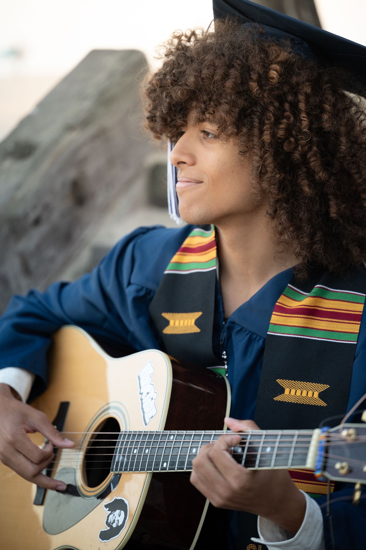 Graduate with curly hair plays acoustic guitar wearing cap, gown, and colorful kente stole at outdoor setting.