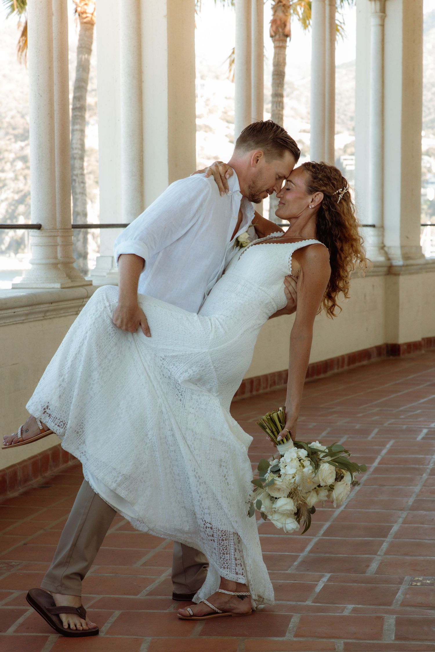 Bride and groom share a romantic dip kiss on a sunlit colonnade, bride holding white bouquet in wedding attire.