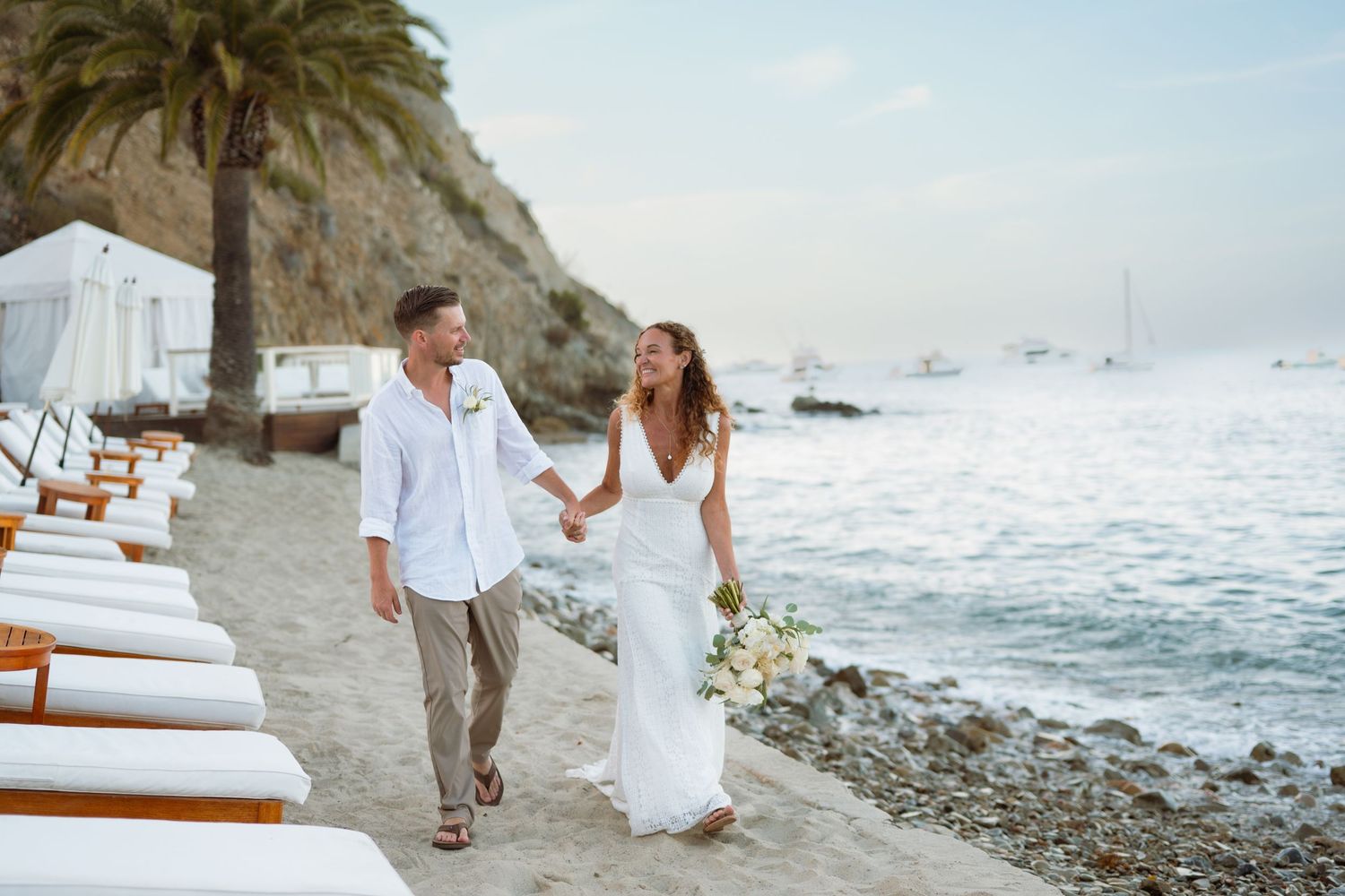 Bride and groom walk hand-in-hand along a scenic beach at sunset with sailboats in background.