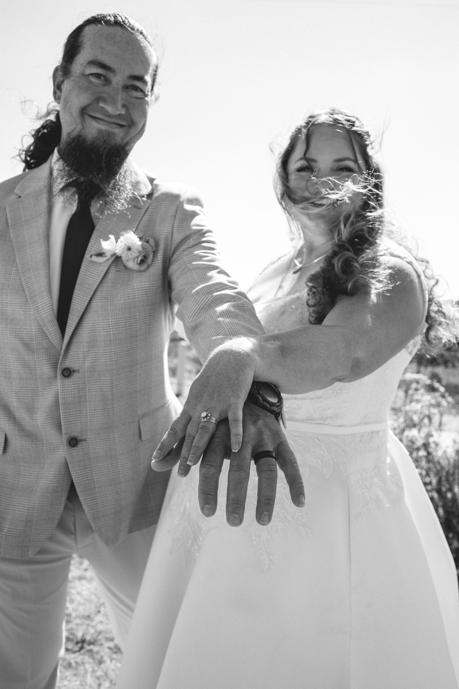 Newlyweds smiling and showing off their wedding rings in a black and white outdoor photo.
