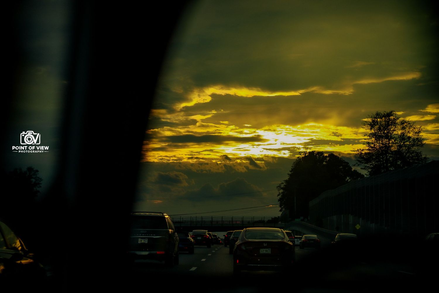 Cars in heavy traffic on a highway at sunset with dramatic golden clouds filling the sky, viewed from inside a vehicle.