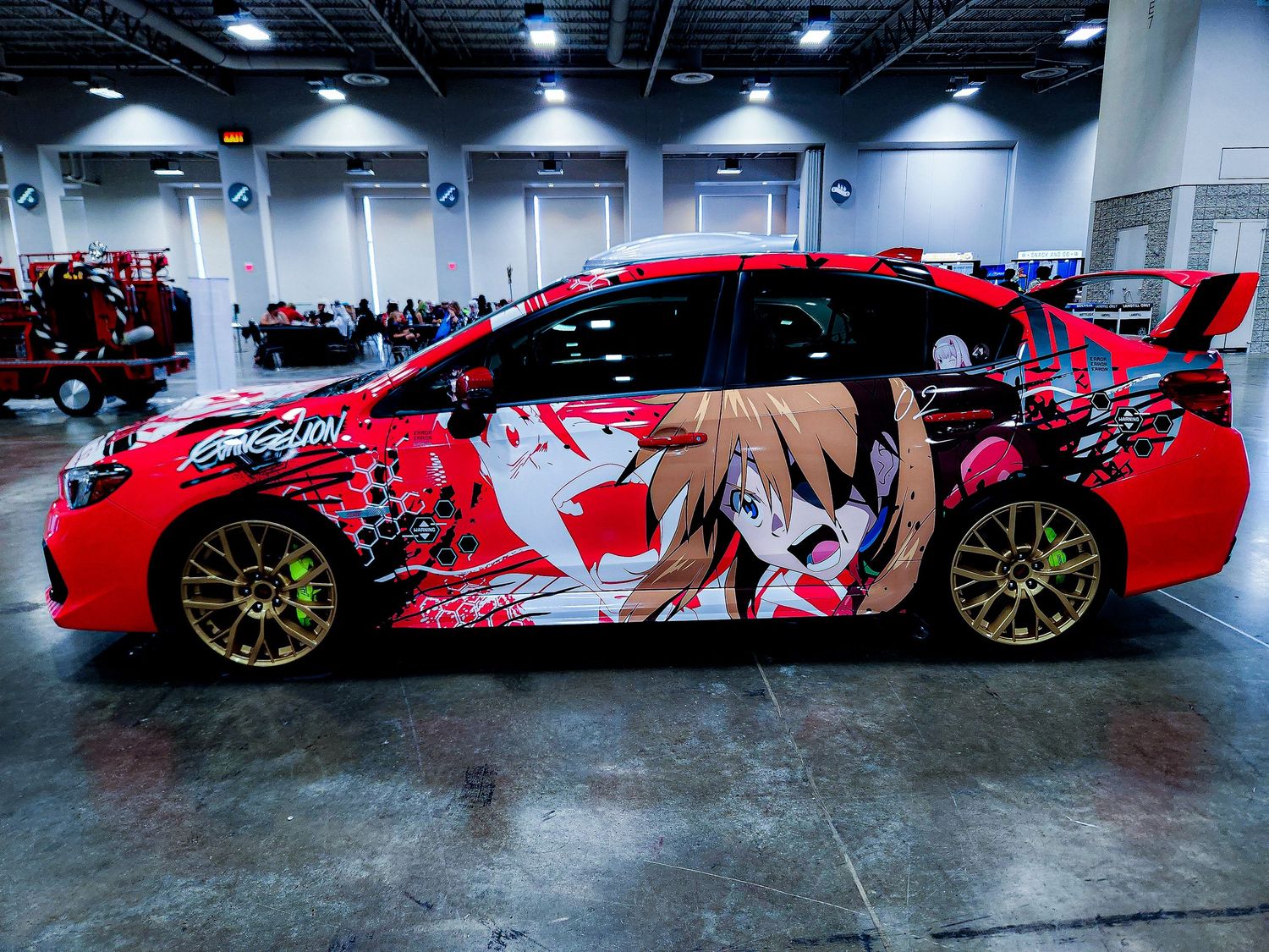 Red Subaru WRX STI with Evangelion anime vinyl wrap featuring blonde character at car show.