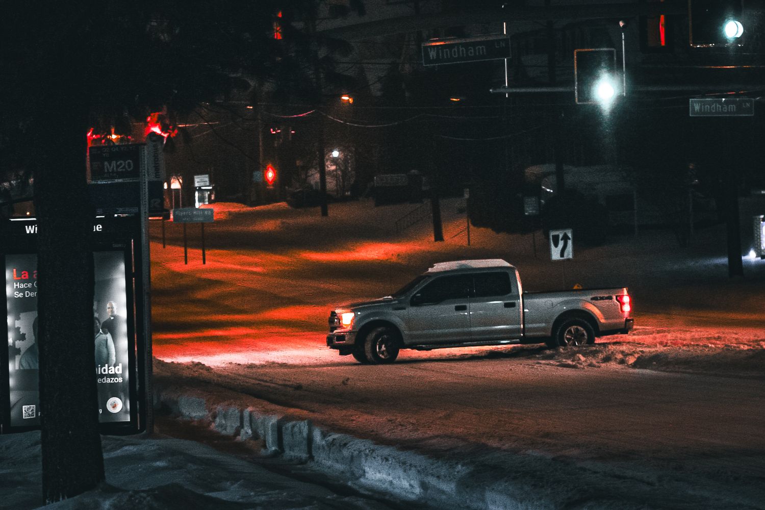 Pickup truck parked at snowy urban intersection illuminated by red street lights at night.