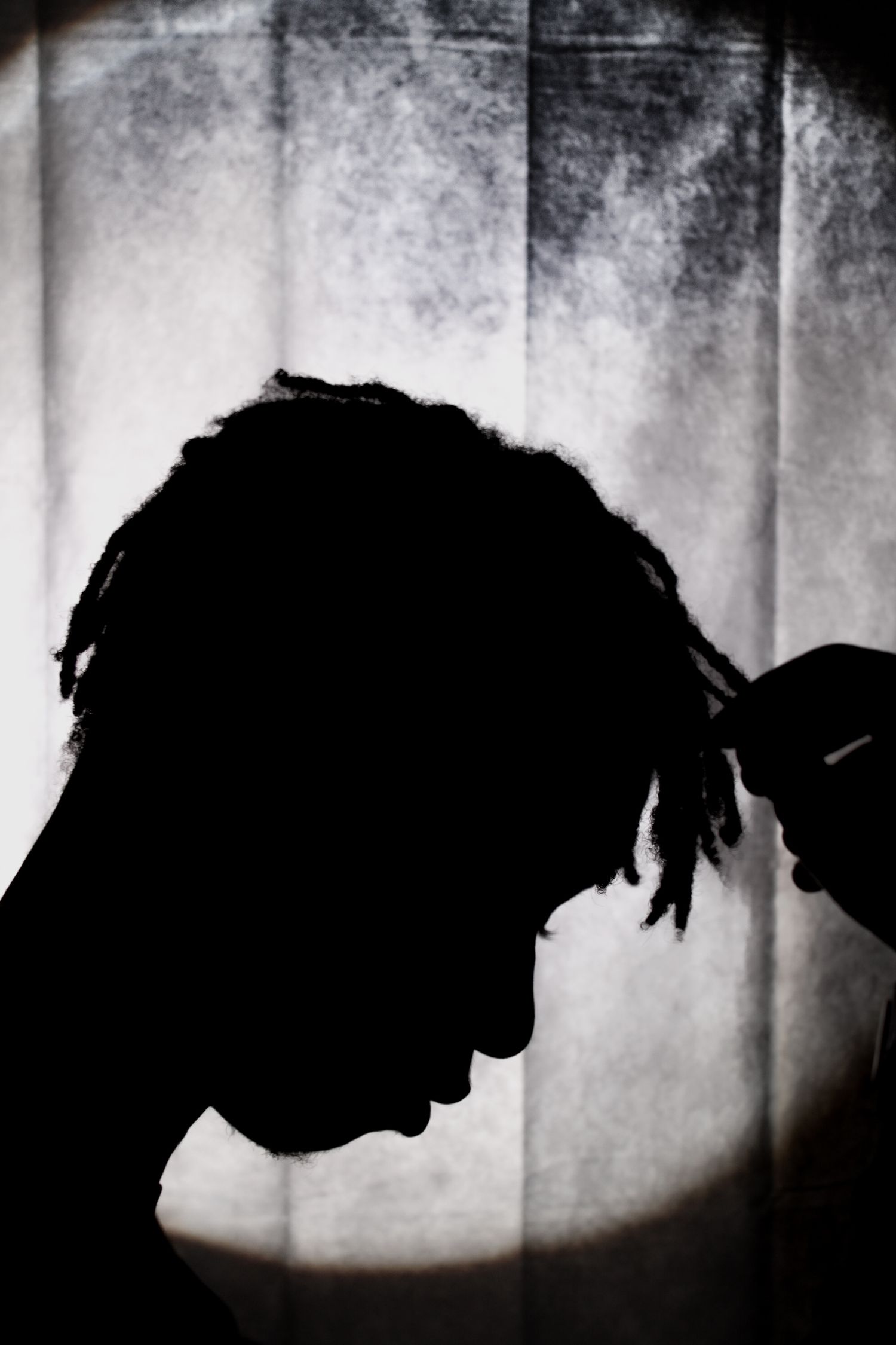 A dramatic silhouette profile of a person with dreadlocks holding their head against a textured gray background.