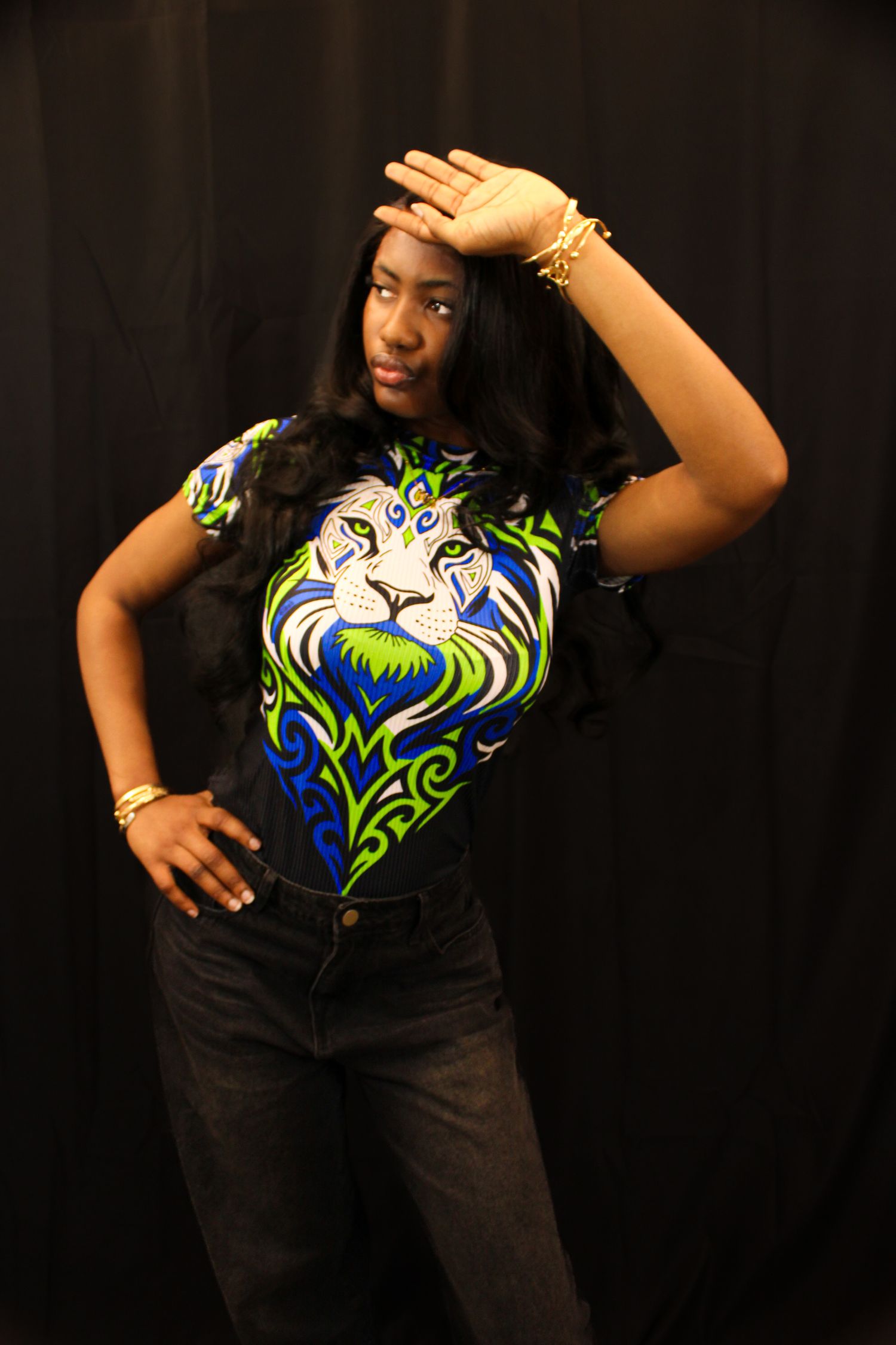 Woman in bold lion-print shirt and black jeans posing with hand raised against dark background.