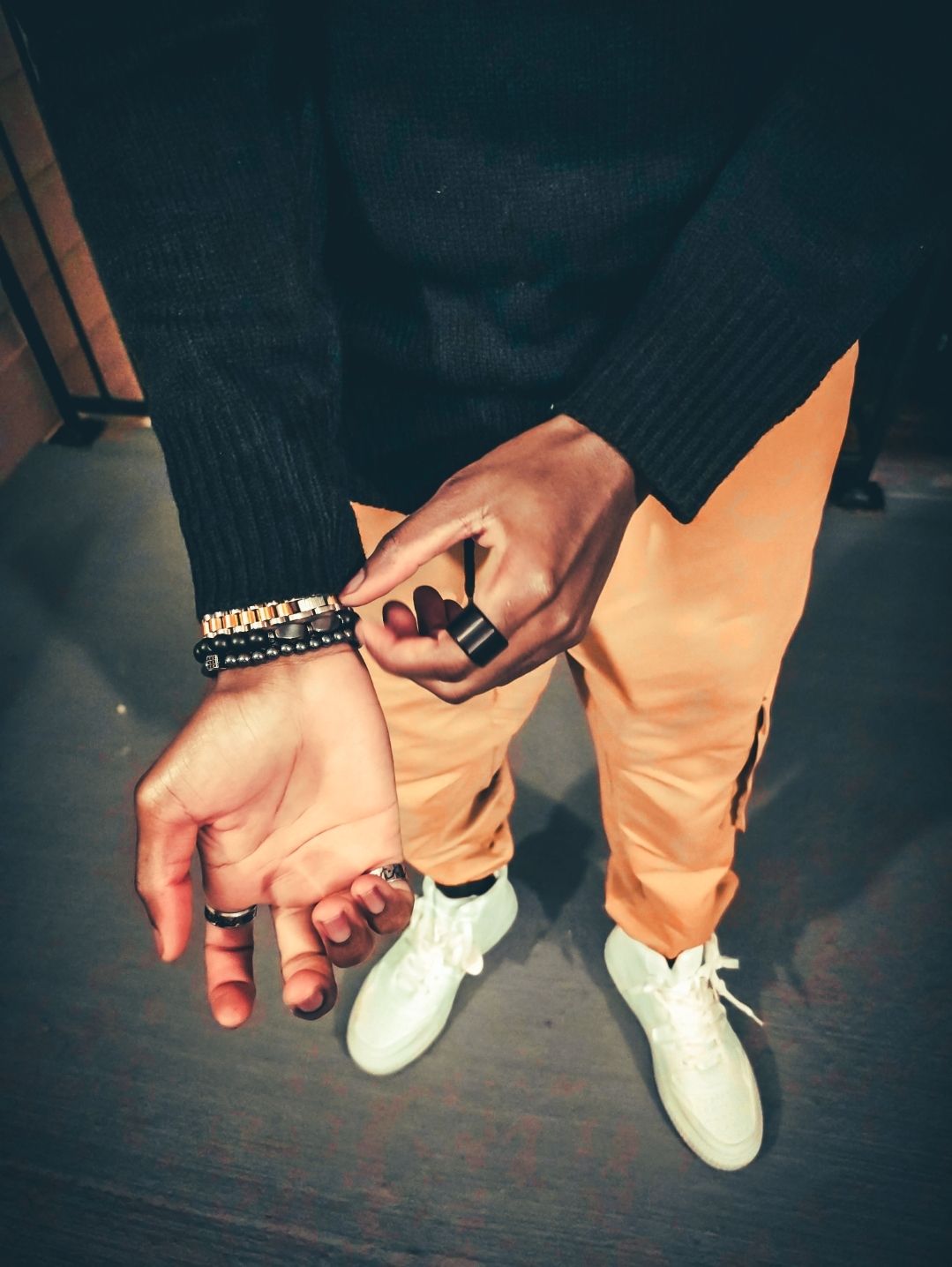 Close-up of hands with rings and bracelets wearing orange pants and white sneakers indoors.