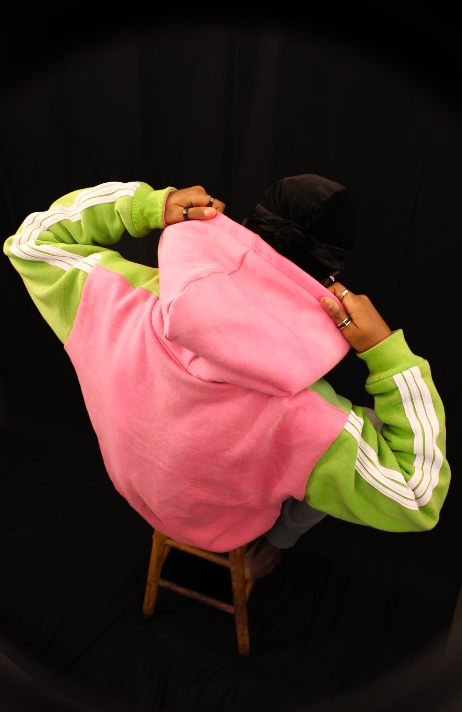 Overhead view of a person in a pink and green Adidas hoodie sitting on a wooden stool against black background.