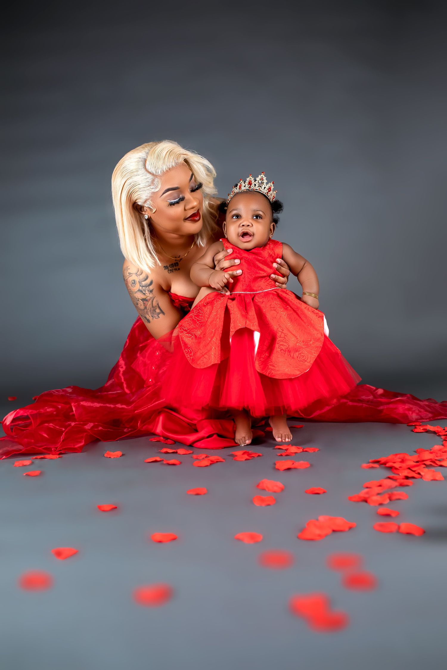 Mother in red dress poses with baby wearing crown and matching red tulle dress on studio floor.