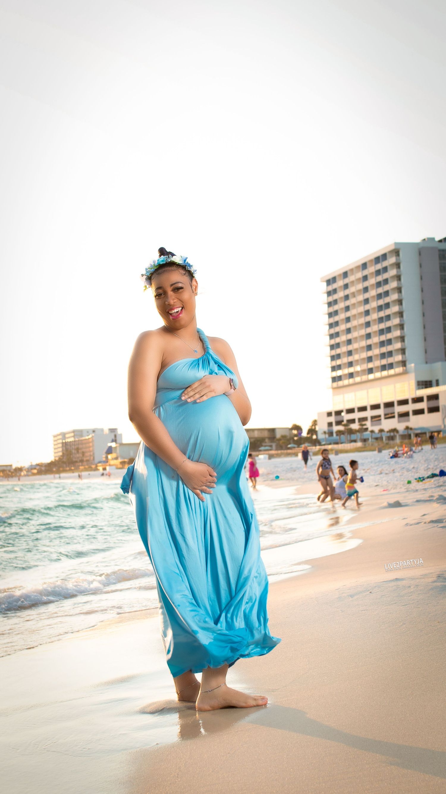 Pregnant woman in flowing blue dress holding belly on sunny beach with ocean waves and resort buildings.