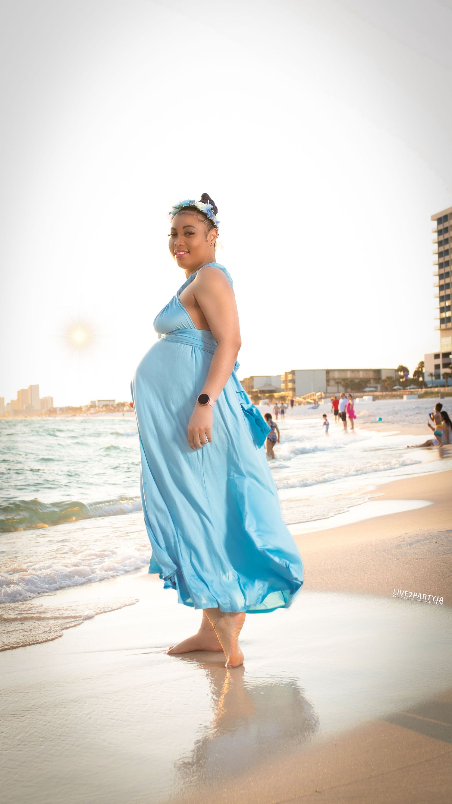 Pregnant woman in turquoise dress walking barefoot on beach shoreline with wind blowing her gown.
