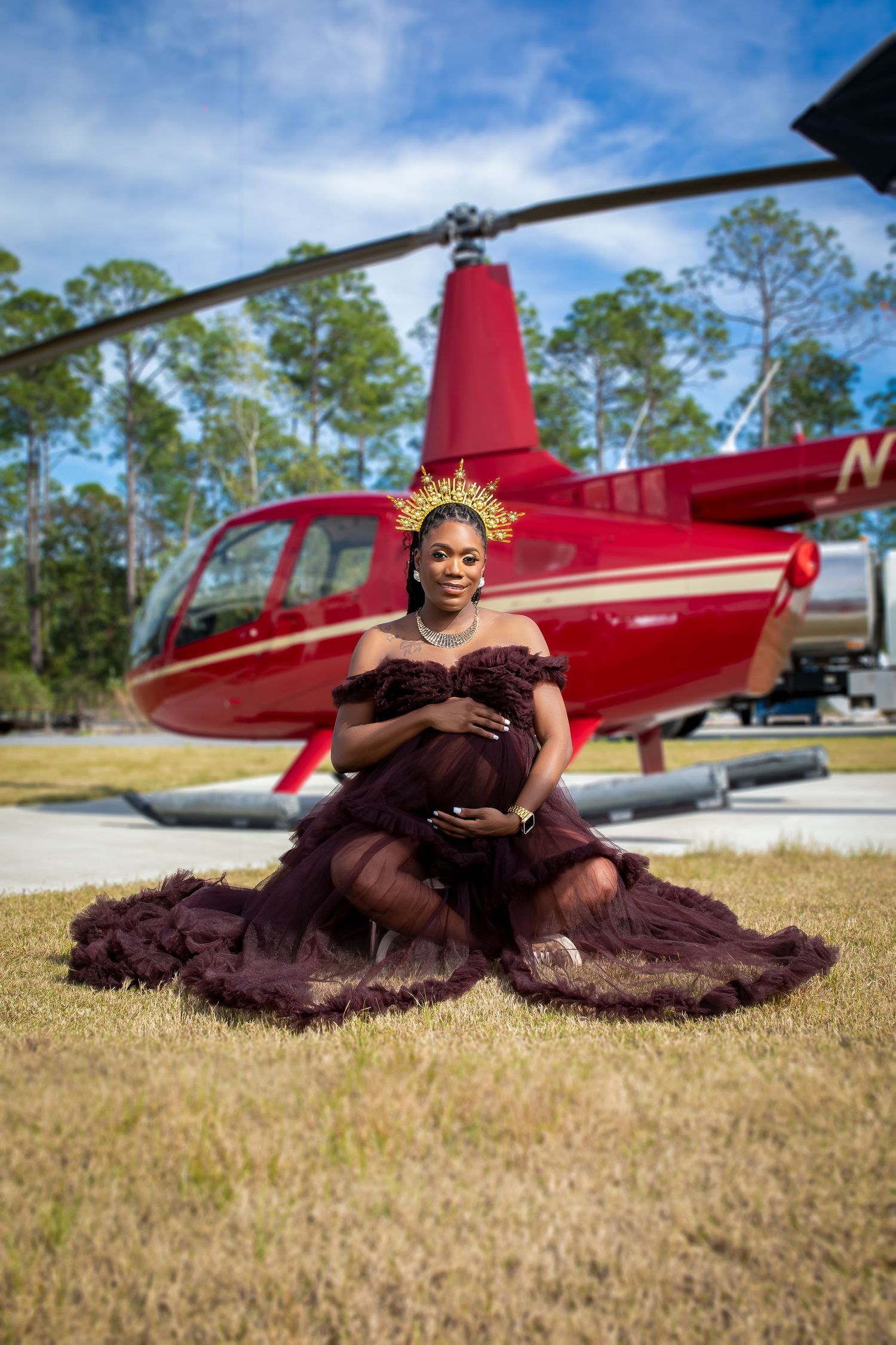 Pregnant woman in dark burgundy tulle gown with gold crown seated gracefully before red helicopter.
