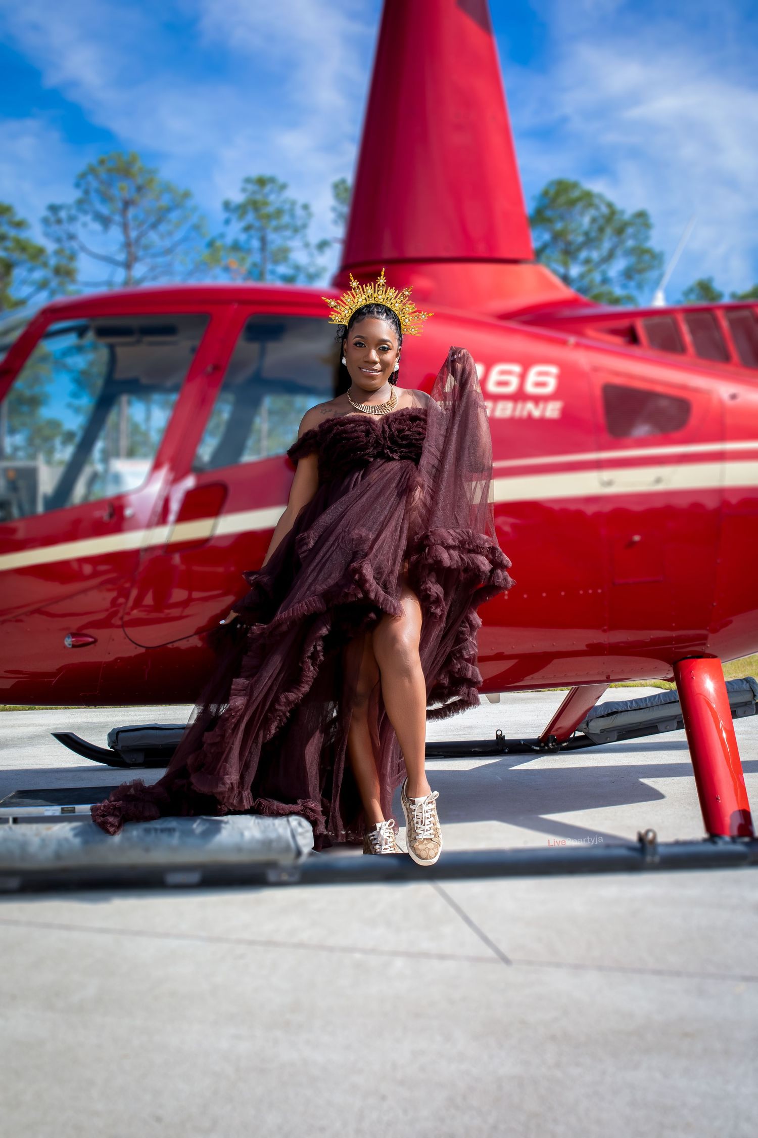 Pregnant woman in maroon ruffled gown with gold crown standing on red helicopter skid outdoors.